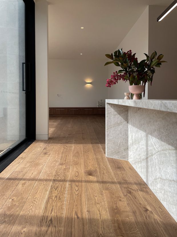 A refined open-plan interior viewed from a large black-framed glazed entry, with warm timber flooring, a long dark timber built-in cabinet, and a sculptural pale stone island or counter in the foreground. Neutral plaster walls, recessed lighting, a linear pendant, and a large floral arrangement create a calm, contemporary atmosphere.
