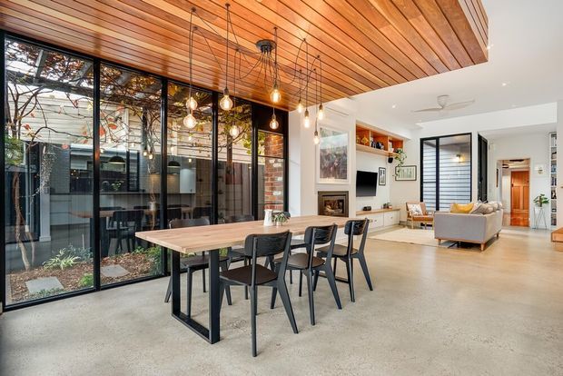Open-plan contemporary dining and living area with a long timber dining table and black metal-framed chairs. The space features a dramatic slatted timber ceiling with hanging pendant lights, polished concrete flooring, expansive black-framed glazing overlooking a planted courtyard, exposed brick detailing, and a connected lounge area with built-in shelving and a fireplace.