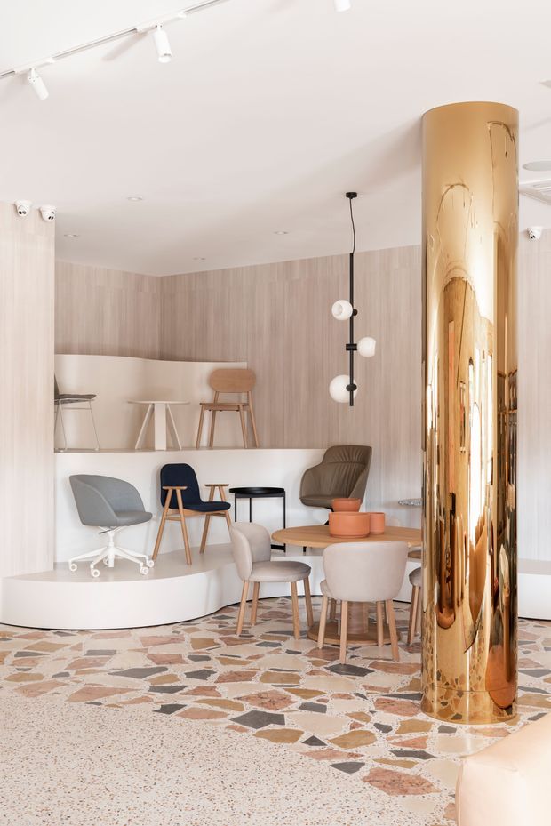 A bright, contemporary commercial interior with a reception or lounge setting. The space features pale timber-look wall panelling, white walls and ceiling, a raised curved platform with assorted lounge and task chairs, small round tables, sculptural pendant lighting, and a striking polished gold cylindrical column. The floor combines terrazzo with an irregular patterned stone or terrazzo inlay.