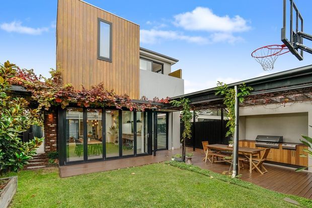 A contemporary residential courtyard viewed from the garden, featuring a two-storey extension with warm vertical timber cladding, exposed brickwork, extensive black-framed glazing, and a covered outdoor entertaining area. The courtyard includes timber decking, a dining setting, built-in outdoor cabinetry with a barbecue, climbing vines, and a basketball hoop. A white upper-level volume and a partially visible interior add to the modern composition.