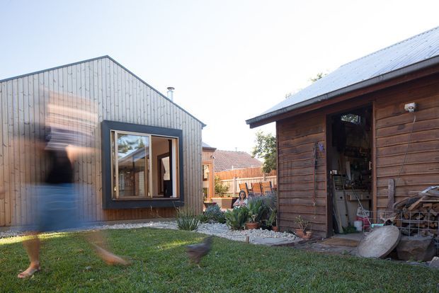 A contemporary backyard scene with a compact grey-clad building featuring a steeply pitched roof and dark-framed window, alongside a rustic timber shed or workshop with a corrugated metal roof and open storage area. A landscaped gravel garden connects the structures, with blurred people moving across the lawn.
