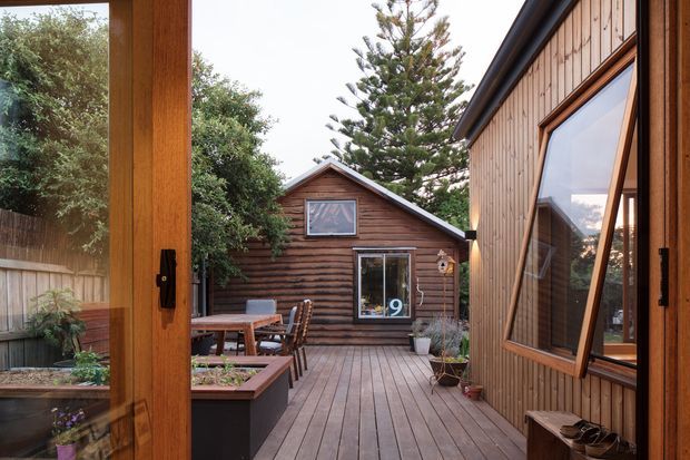 View from inside a timber-framed building onto a compact landscaped courtyard and timber deck. The space is enclosed by vertical timber-clad walls and a small timber outbuilding with a pitched roof. Outdoor dining furniture, raised garden beds, potted plants, and large timber-framed glazing surround the deck, creating a warm, private residential courtyard setting.