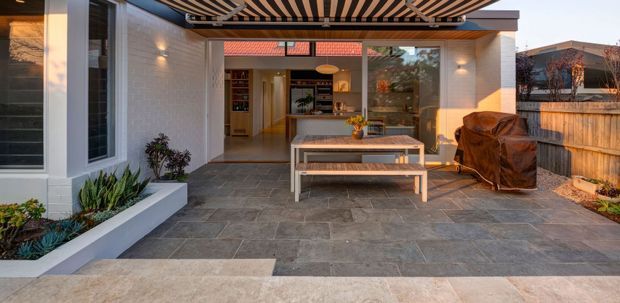 A contemporary covered outdoor entertaining area with a paved terrace, built-in garden beds, dining table and bench seating, and a covered barbecue. The space opens directly into a bright interior kitchen or living area through large glazed doors. A timber pergola with striped slatted screening provides shade, while rendered white walls and timber fencing frame the courtyard.