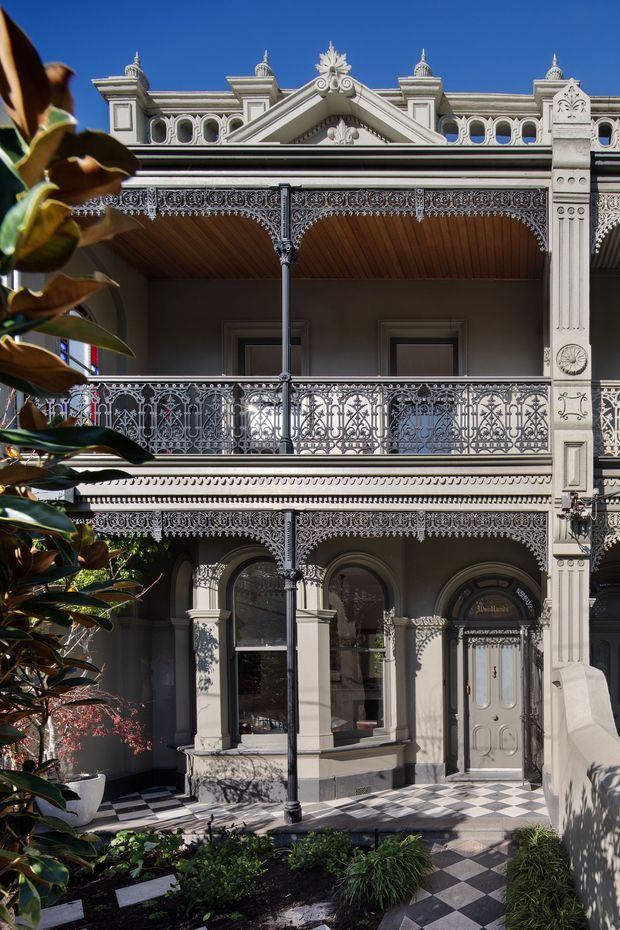 Front facade of an ornate multi-storey Victorian-era terrace or townhouse, featuring elaborate cast-iron lacework balconies and balustrades, decorative cornices and gable detailing, classical columns, arched windows, and a paneled entry door. A black-and-white checkerboard tiled path leads through a landscaped garden, with foliage framing the left side. Bright clear daylight emphasizes the detailed heritage architecture.