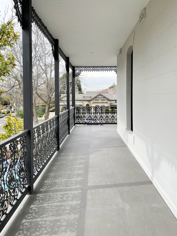 A long, covered upper-level veranda or balcony with a white-lined ceiling, white horizontal-clad wall, large grey floor tiles, and ornate black metal balustrades and posts. Decorative lace-like railing casts patterned shadows across the floor. An arched opening is visible along the wall, with mature trees and neighbouring heritage-style buildings beyond the open side.