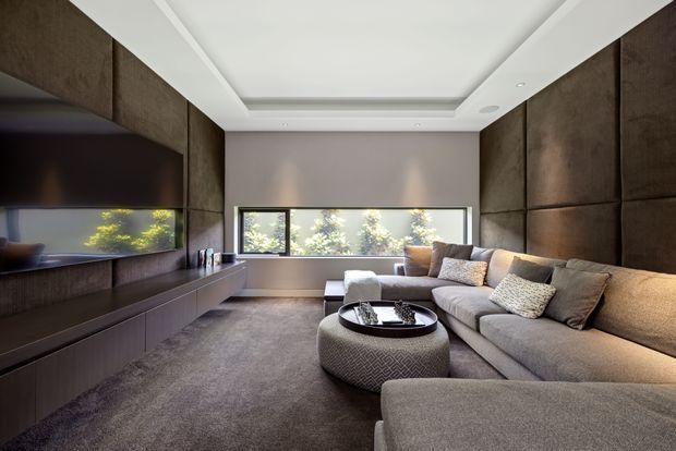 A refined, contemporary media room with a large sectional sofa, upholstered round ottoman, wall-mounted television, and low built-in media cabinetry. Dark textured wall panelling frames the room, while a wide horizontal window provides natural light and views of greenery. The recessed tray ceiling, neutral upholstery, and soft layered lighting create a calm, luxurious atmosphere.