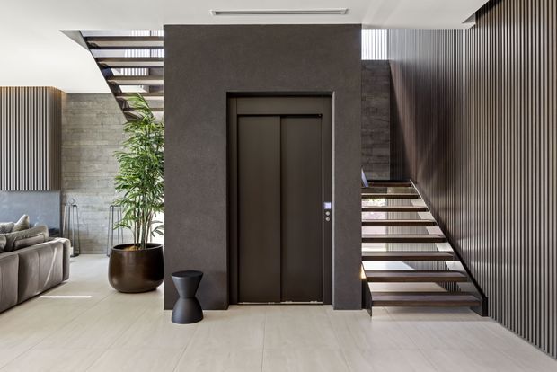 Contemporary interior foyer featuring a dark textured elevator surround, floating timber staircases on both sides, vertical slatted wall paneling, pale large-format floor tiles, and a concrete-look feature wall. A partial living area with sofa and indoor planter is visible to the left.