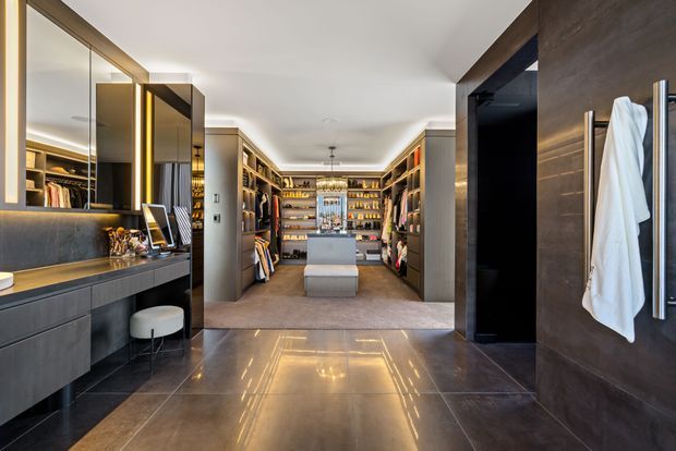 Luxurious walk-in wardrobe and dressing room viewed from a dark timber-framed entry. The space has extensive floor-to-ceiling open shelving and hanging storage, a central island with illuminated glass display cabinetry, a long dark built-in vanity with mirror, stool and lighting, and large polished stone-look floor tiles. Warm integrated lighting creates a refined, high-end atmosphere.