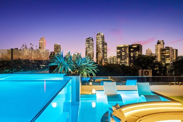 A dusk rooftop pool area overlooking a brightly lit urban skyline. The scene features a glowing turquoise swimming pool, illuminated submerged and poolside seating, sculptural curved lounge furniture, glass balustrades, and spiky architectural planting. The atmosphere is vibrant, luxurious, and contemporary.