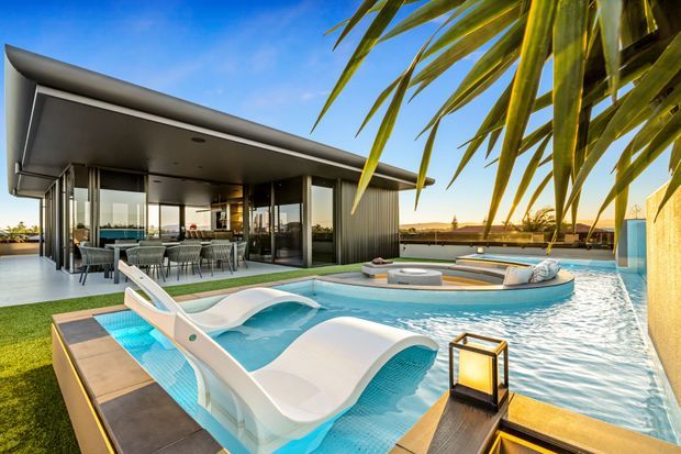 A contemporary single-storey pavilion-style home opens onto a resort-like pool terrace. The image features a sculptural turquoise swimming pool with integrated white loungers, a timber-edged deck, outdoor lantern, and a covered dining area behind full-height glazing. The flat-roofed structure uses dark metal framing, extensive glass, and ribbed vertical cladding, with palm foliage and a low sunset horizon in the background.