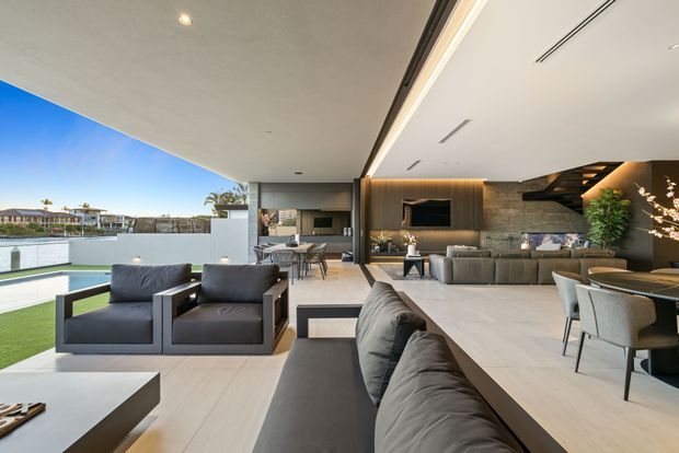 A spacious contemporary open-plan living area with a large, fully opened glazed wall connecting the interior to an outdoor terrace and waterfront setting. The room includes a grey sectional sofa, matching lounge chairs, a low coffee table, dining furniture, and a compact kitchen or bar area in the background. Warm timber wall panelling frames the television, while a dark staircase and concealed linear lighting add architectural definition. Pale flooring, neutral furnishings, and abundant daylight create a refined indoor-outdoor atmosphere.