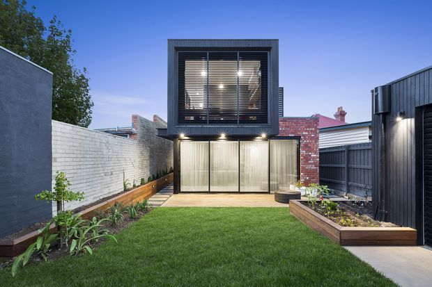 A compact contemporary urban backyard with a newly built two-storey structure at the rear. The upper level has dark metal cladding, large framed glazing and horizontal blinds, while the lower level features translucent panelled doors and downlights. Red brick and weatherboard structures surround a neatly landscaped lawn with raised timber garden beds, boundary walls and a paved side path.