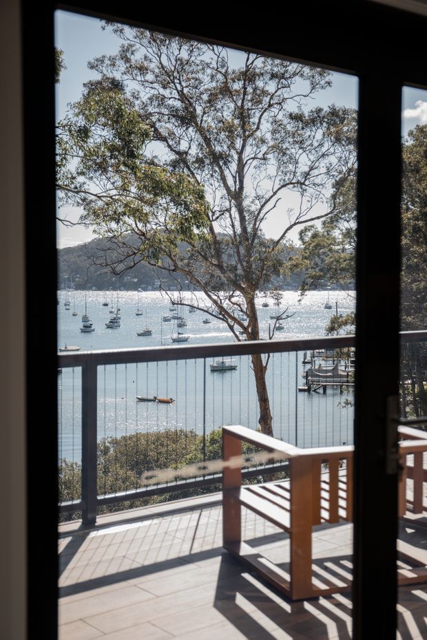 A view from inside through large glazed doors onto a timber-clad balcony or terrace overlooking a sheltered, boat-filled bay. The terrace has dark grey tiled or concrete flooring, timber handrails, and dark metal balustrades with vertical cables. Native trees frame the water and wooded shoreline beyond. Strong sunlight creates crisp shadows across the deck.