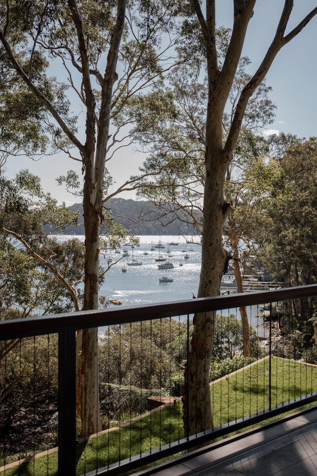 A view from a residential deck or balcony through tall eucalyptus trees toward a sunlit bay filled with anchored boats. The foreground features a dark metal balustrade with fine vertical cables, timber-toned decking, and a sloping landscaped lawn below. Forested hills and waterfront scenery form the background.