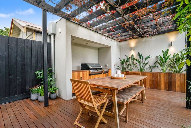 A landscaped outdoor dining and entertaining area on a timber deck, featuring a built-in barbecue, timber dining table and chairs, raised planter boxes with lush greenery, and a dark pergola structure overhead covered with climbing vines. White rendered walls and warm timber finishes create a sheltered courtyard-like setting.