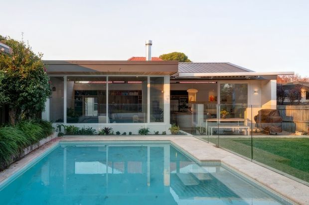 Single-storey contemporary home viewed from the backyard, with a large swimming pool in the foreground, broad glazed living spaces, a covered outdoor entertaining area, and a low-pitched metal roof. The building uses a restrained palette of white walls, dark grey roofing, and extensive glass, with landscaped planting and a timber privacy fence around the yard.