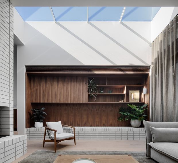 Bright contemporary living room with a double-height skylit ceiling, white tiled fireplace or feature wall at left, and a large built-in dark timber display and seating wall. A low white tiled plinth runs beneath the joinery, with a woven timber armchair, grey sofa, plants, and floor-to-ceiling curtains creating a warm refined interior.