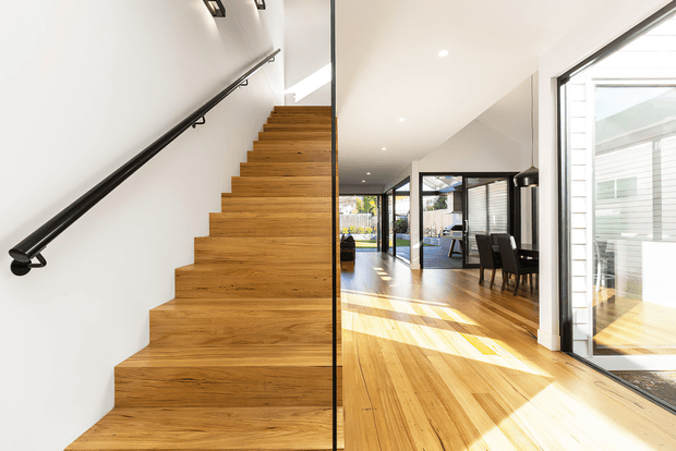 Bright contemporary open-plan interior viewed from the base of a timber staircase. The staircase has broad solid-wood treads with a minimalist black handrail and a clear glass balustrade. Light timber flooring continues into the dining and living area, which is defined by extensive black-framed glazing and strong natural sunlight. White walls and ceiling create an airy, clean composition.