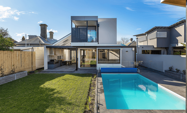 Exterior rear view of a contemporary multi-level residence with a compact rectangular swimming pool, paved pool terrace, lawn, timber boundary fencing, and extensive glazing. The home combines a light rendered lower level with a dark upper volume featuring horizontal screening and a projecting balcony or window enclosure. A covered outdoor kitchen or entertaining area is visible beneath the ground-floor overhang. Traditional neighbouring roof forms contrast with the modern addition.
