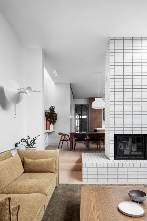 A bright, refined open-plan living and dining space with a neutral palette. The foreground contains a large textured beige sectional sofa and timber coffee table, while the background features a dining table with dark chairs, a timber-toned built-in wall, and a black-framed glazed doorway. A prominent white brick-tiled fireplace and hearth divides the living and dining zones. White walls, pale timber flooring, sculptural wall lighting, indoor plants, and simple contemporary furnishings create a calm, warm interior.