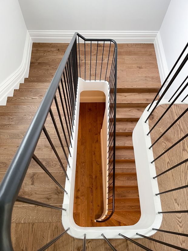 Top-down view of a bright, multi-level stairwell with warm timber stair treads and flooring, white walls and curved white stair edges, detailed white crown moulding, and slim black metal balustrades surrounding the central void.