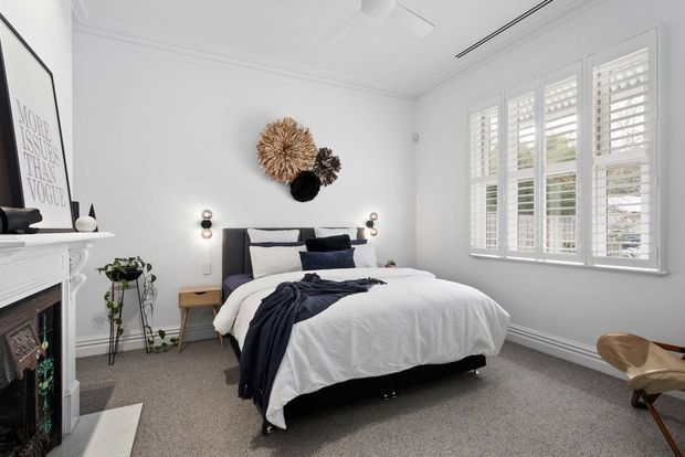 Bright, refined bedroom with a layered white bed, dark upholstered headboard, navy and charcoal textiles, pale grey carpet, and white walls with decorative cornices and skirting. Plantation shutters cover the large windows, while a traditional white fireplace, simple bedside tables, wall sconces, and sculptural woven wall decor add character. The room has a balanced contemporary-traditional feel with abundant natural light.