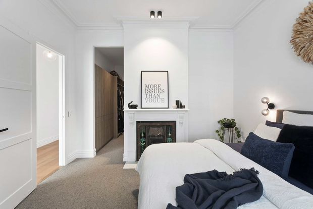Bright, refined bedroom with white panelled walls, ornate ceiling cornices, a classic mantel fireplace, and a large upholstered bed dressed in white linen with navy cushions. A walk-in wardrobe or dressing area is visible through the open doorway, with warm timber joinery. The room combines traditional architectural detailing with contemporary furnishings and soft neutral tones.