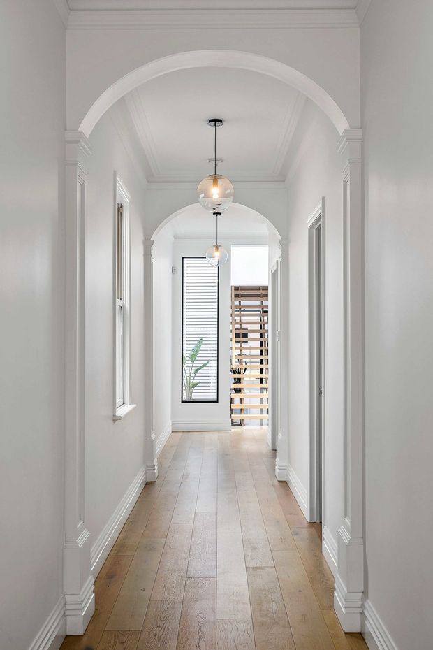 Bright, elegant residential hallway viewed through a sequence of white decorative archways. The corridor has white painted walls, ornate wall and ceiling mouldings, white door and window trim, pale timber plank flooring, and two suspended spherical pendant lights. A tall narrow window at the far end and a timber slatted screen bring daylight into the space.