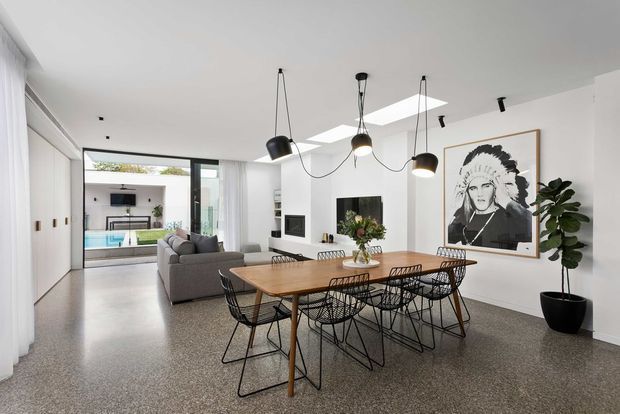 Bright open-plan contemporary dining and living area with polished terrazzo flooring, white walls, extensive glazing, skylights, a timber dining table with black wire-frame chairs, a grey sofa, minimalist pendant lights, built-in cabinetry, and a large monochrome artwork. A courtyard and pool are visible beyond the glazing.