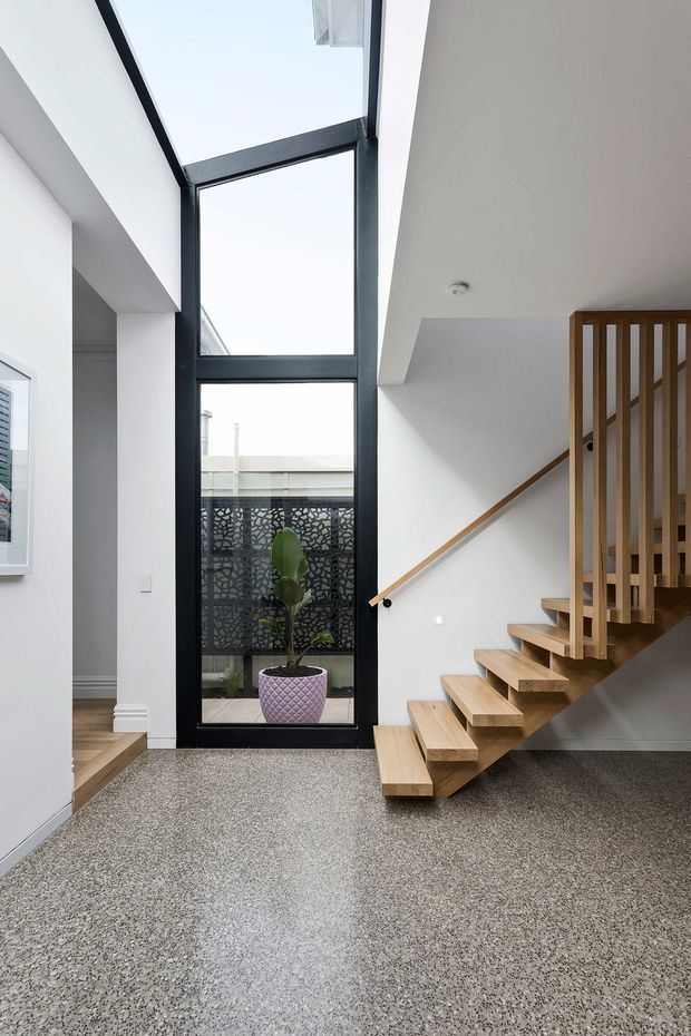 Bright contemporary residential stair hall with a tall black-framed glazed opening, white walls, a floating timber staircase with matching vertical balustrade, and a speckled polished floor. A small planted courtyard is visible through the glazing, while the open stair and high vertical glazing create an airy, light-filled entry space.
