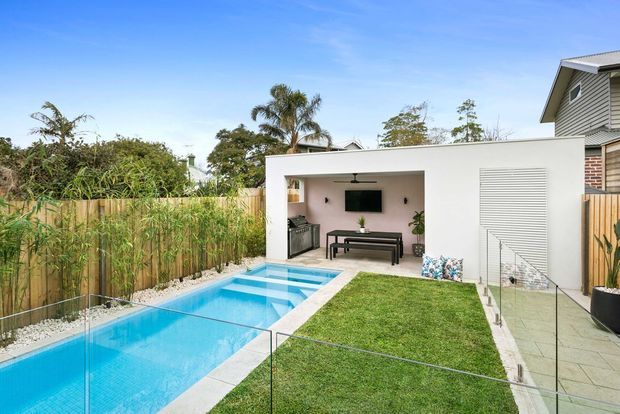 A bright contemporary backyard featuring a long rectangular swimming pool, manicured lawn, glass pool fencing, and a covered outdoor entertaining pavilion. The pavilion has a flat white roof, built-in outdoor kitchen or barbecue cabinetry, ceiling fan, wall-mounted television, and outdoor lounge furniture. Timber fencing and tropical planting frame the residential setting.