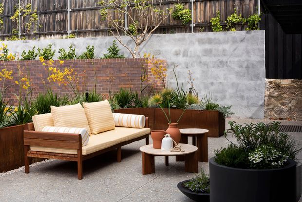 A landscaped urban courtyard or outdoor lounge with a mid-century-style timber sofa and matching striped cushions, layered round coffee tables, and abundant potted and garden planting. The space is enclosed by weathered timber fencing, rendered concrete walls, a red brick retaining wall, and raised rust-toned planter beds, creating a warm, textural contemporary garden setting.
