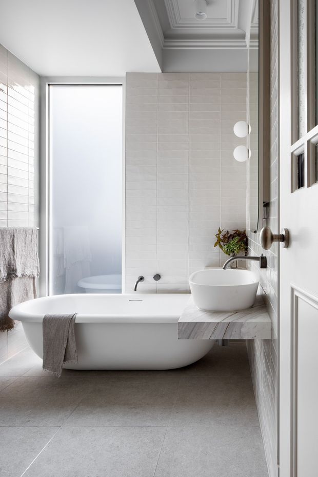 A bright, refined bathroom featuring a freestanding white bathtub, wall-mounted basin with a pale stone or marble benchtop, white rectangular wall tiles, large grey floor tiles, and a frosted-glass shower enclosure. Traditional decorative ceiling mouldings and a white paneled door contrast with the clean contemporary fixtures. Soft grey textiles and subtle greenery add warmth.