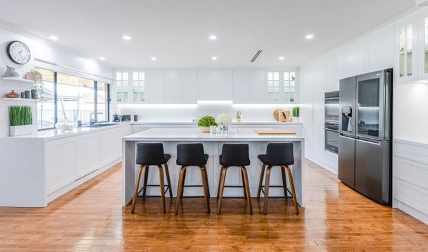 Bright, spacious white kitchen with extensive built-in cabinetry, a large central island with seating for four, stainless-steel appliances, recessed lighting, and warm timber flooring. Glazed doors and windows provide abundant natural light, while dark bar stools create contrast.