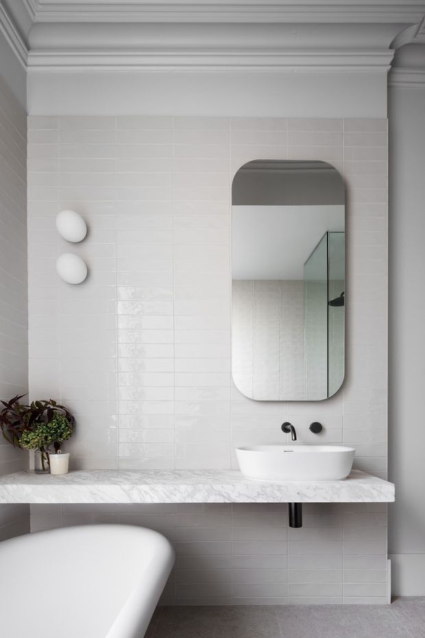 Refined contemporary bathroom with a freestanding bathtub in the foreground, a wall-mounted white basin, black tapware, rounded rectangular mirror, glossy white tiled walls, and a pale marble-look vanity shelf. Decorative moulded cornice and wall detailing add a classic architectural touch, while a glass shower screen is reflected in the mirror.