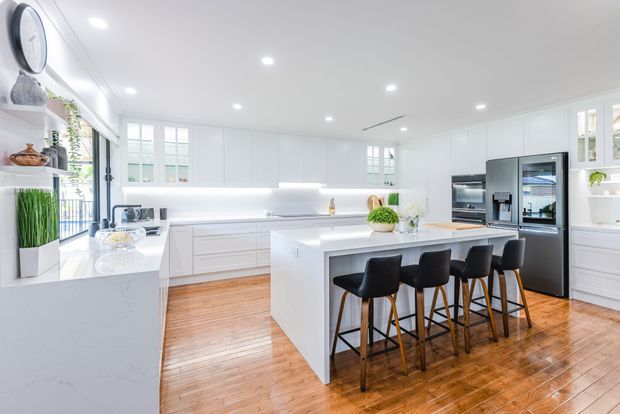 A bright, spacious contemporary white kitchen with extensive built-in cabinetry, a large central island with seating for five, integrated appliances, and warm polished timber flooring. White shaker-style cabinetry, recessed ceiling downlights, and large glazed windows create an airy, high-contrast interior.