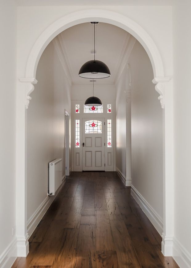 A narrow, elegant residential hallway viewed through a large white decorative arched opening. The corridor has dark timber flooring, white painted walls with ornate moulded architraves and skirting, a radiator, and a traditional panelled front door with distinctive leadlight glazing featuring red floral motifs. Two black dome pendant lights hang centrally, creating a strong contrast with the light interior.