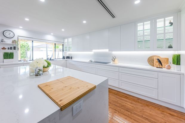 Bright, spacious white kitchen with extensive shaker-style cabinetry, a large white stone island, integrated under-cabinet lighting, timber flooring, and a timber chopping board. The room includes a wall of cabinetry, a glazed window with plantation shutters, recessed ceiling lights, and a view toward an outdoor area through the rear glazing.