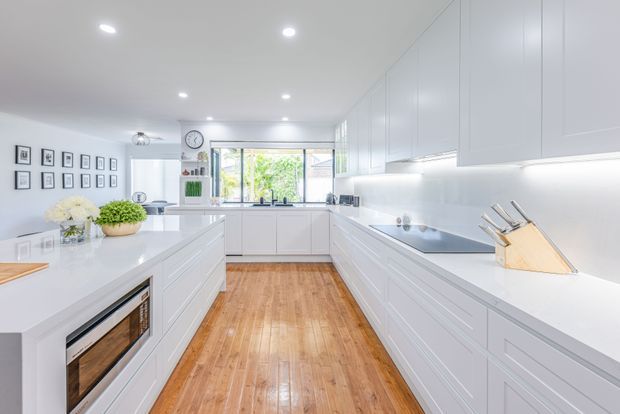 Bright, spacious contemporary white kitchen with extensive floor-to-ceiling cabinetry, a large central island with integrated appliance, pale benchtops, under-cabinet lighting, and timber flooring. The room opens toward a windowed rear work area and includes a small dining or display zone with framed artwork.