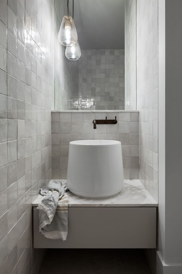 A compact, narrow bathroom featuring full-height pale grey square wall tiles, a floating dark grey vanity with a light stone benchtop, and a large white vessel basin. A dark wall-mounted tap and two clear glass pendant lights provide contrast, while a subtly varied square-tile feature wall is visible behind the basin. The space has a restrained, contemporary minimalist appearance with cool neutral tones.