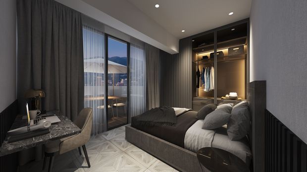 A sophisticated contemporary bedroom with a dark upholstered bed, layered grey bedding, full-height curtains, patterned light flooring, and a built-in illuminated wardrobe. A compact desk and chair sit beside floor-to-ceiling glazing, which opens toward a balcony or exterior view. Recessed ceiling lights and warm wardrobe lighting create a refined, moody atmosphere.