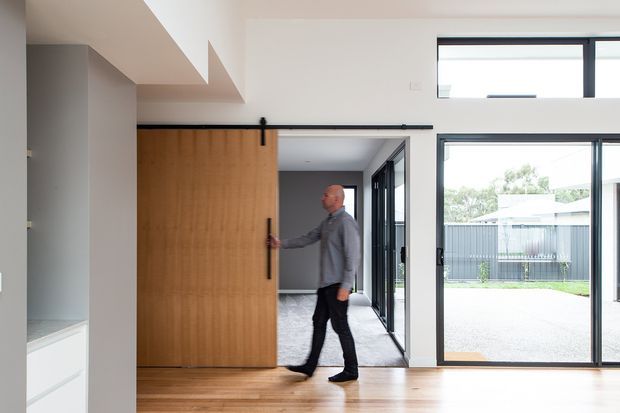 Bright contemporary interior showing a large timber sliding barn-style door on black hardware, opening from a hallway or living area toward a carpeted room. The space has pale walls, light timber flooring, large black-framed glazed doors and windows, and a clean minimal fit-out. A fenced outdoor area is visible through the glazing.