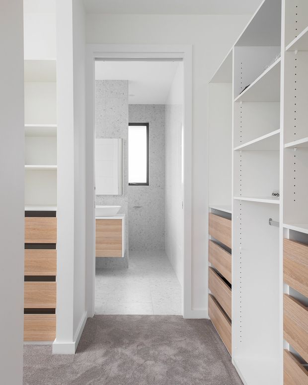 Bright, compact walk-in wardrobe with white open shelving and light timber drawer fronts on both sides, opening directly into a modern bathroom. The bathroom features pale grey tiled walls and floor, a white vanity basin, and a narrow black-framed window. Soft grey carpet covers the wardrobe floor, creating a clean, minimalist built-in storage space.
