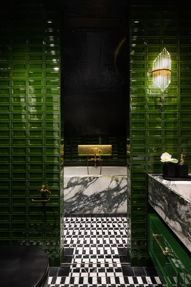 A dramatic, highly detailed bathroom interior with glossy deep-green wall tiles, a black-and-white geometric tiled floor, and a freestanding marble-clad bathtub positioned beneath a dark shower wall. The space includes dark green built-in cabinetry, heavily veined marble surfaces, brass plumbing fixtures, a sculptural pendant light, and a black toilet partially visible in the foreground. The overall design is luxurious, dark, and Art Deco-inspired.