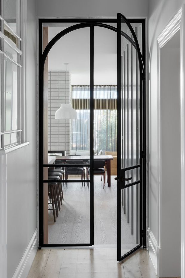 View down a bright, finely trimmed hallway through a black metal-framed glazed arched door into a dining area. The door has a large curved arch, divided glass panels, and is shown partly open. Beyond it are a timber dining table, dark dining chairs, a white pendant light, pale timber flooring, and full-height windows with sheer curtains. White painted walls, decorative skirting, and architectural mouldings frame the passage.