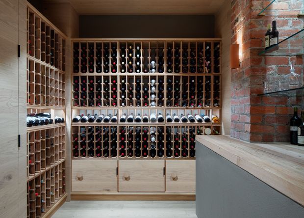 A compact, built-in wine storage room with extensive timber wine racks covering the walls and holding numerous bottles. Pale timber cabinetry and drawers line the lower section, while a substantial exposed red-brick wall and timber-topped counter create a warm, industrial character. The space is viewed directly from within the cellar, with additional bottle storage visible along the left wall.
