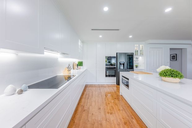 Bright, spacious white kitchen with extensive built-in cabinetry, a long stone-look benchtop, integrated cooktop and appliances, and a central island with timber breakfast-bar detailing. Warm timber flooring contrasts with the predominantly white fit-out, while recessed ceiling lights and under-cabinet lighting create an airy, clean atmosphere.