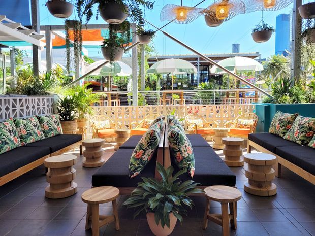 Bright, tropical-inspired outdoor lounge or hospitality terrace with dark built-in sofas, patterned botanical cushions, low timber stools and tables, hanging planters, decorative pendant lights, and a white geometric breeze-block balustrade. The open-air space overlooks a lively landscaped courtyard with umbrellas and additional seating.