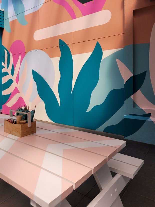 A vibrant outdoor or semi-enclosed dining area featuring a pale pink picnic-style table and benches against a large abstract botanical mural. The mural uses bold teal, pink, peach, cream, and dark blue organic shapes, creating a playful contemporary setting. Dark tiled flooring and a small wooden container with plants or utensils are also visible.