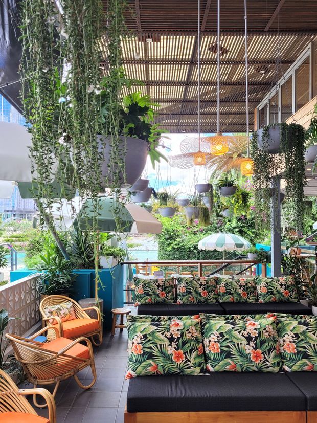 A lush, tropical-style outdoor lounge or café terrace beneath a slatted timber pergola. The space is densely decorated with hanging plants, green walls, and suspended woven pendant lights. Black seating with vibrant botanical floral upholstery forms the main seating area, complemented by rattan lounge chairs with orange cushions. A pool or bright outdoor area is visible beyond the railing.